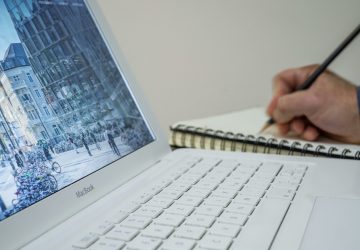 A person taking notes in a notebook beside an open laptop displaying an urban scene.